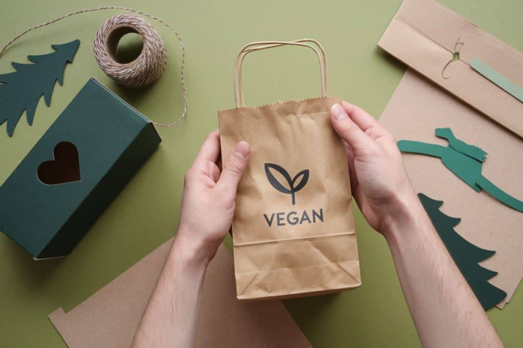 En person håller i en brun papperspåse med texten "VEGAN" tryckt på framsidan. På ett grönt bord ligger ett grönt paket med ett utskuret hjärta, snöre och dekorationer i papper.
