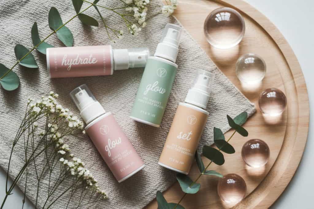 Fyra flaskor setting spray i pastellfärger liggande på tygduk med blommor och glaskulor